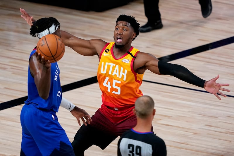 Utah Jazz’s Donovan Mitchell, wearing a gradient city jersey that moves from bright yellow to deep red with the number “45,” has his arms outstretched defending against Denver Nuggets’ Torrey Craig in a blue jersey and a white headband. The back of a referee’s head is at the bottom.