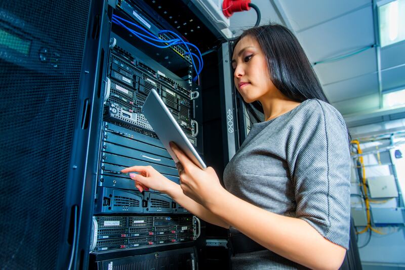 A woman holds a tablet and works in a network server room.