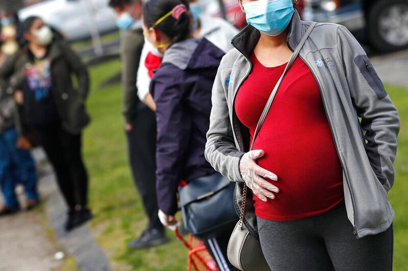 In this Thursday, May 7, 2020 file photo, a pregnant woman wearing a face mask and gloves holds her belly as she waits in line for groceries with hundreds during a food pantry sponsored by Healthy Waltham for those in need due to the COVID-19 virus outbreak, at St. Mary’s Church in Waltham, Mass