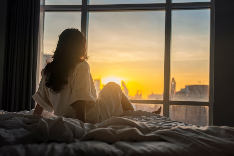 Photo of a woman looking out a bedroom window as the morning sun rises.