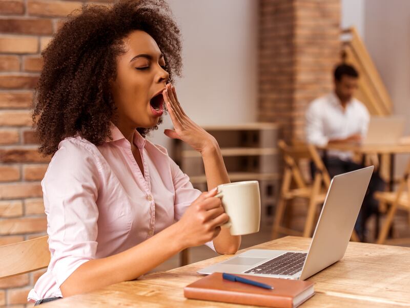 Woman yawning while using a laptop and holding a mug in a cafe.