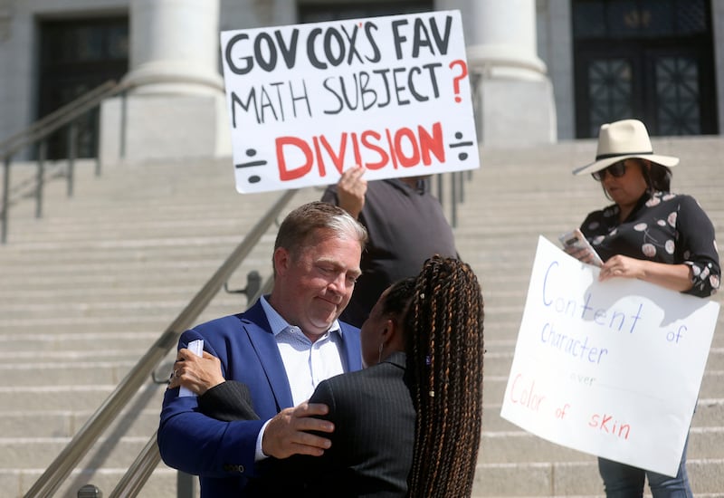 Curtis Linton and Jackie Thompson hug as community activists and educators protest a Utah State Legislature resolution to ban Critical Race Theory concepts. Counterprotestors holding signs stand behind them.
