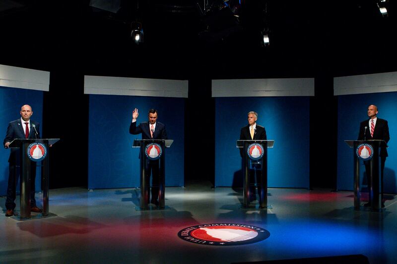 Four men in suits stand in front of equally spaced podiums on a stage with red, white and blue colors on the floor. One man on the middle-left has his hand raised to speak.