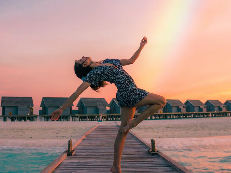 A woman dances on a pier at sunset with a rainbow behind her.