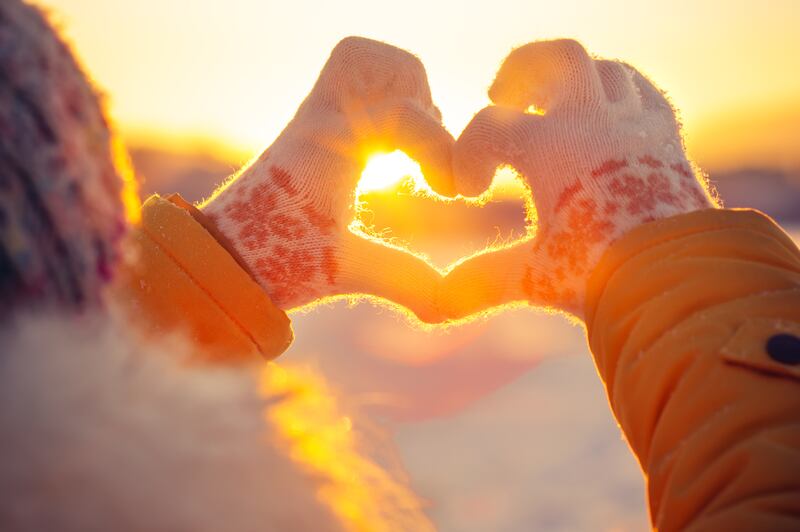 Woman’s hands in winter gloves make a heart symbol with sunset shining through.