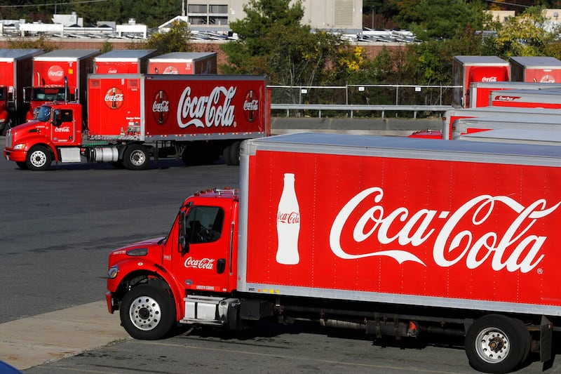 In this photo a truck with the Coca-Cola logo, behind left, maneuvers in a parking lot at a bottling plant in Needham, Mass.