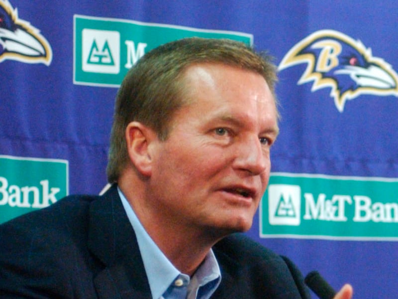Coach Jim Fassel speaks at a press conference. He is in front of a Baltimore Ravens backdrop, wearing a suit and pointing with his index finger.