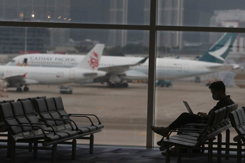 A man sits by himself by a row of chairs at an airport gate. Planes are visible outside the large windows. The planes read “Cathay Pacific.”