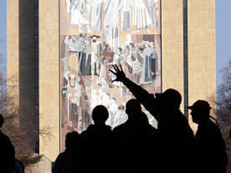 Fans and tourists stand in an entrance to Notre Dame Stadium and listen to a stadium usher on the Notre Dame campus in South Bend, Ind., Friday. In the background is the Hesburgh Library with a mosiac of Jesus on it. The mural is sometimes called "Touchdo