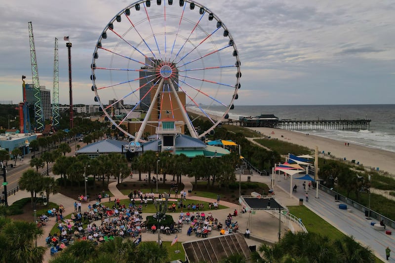 People gather outside next to a large Ferris wheel in an amusement park to watch an outdoor screen.