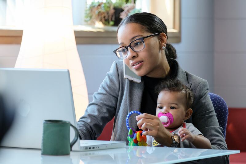 A woman in a suit jacket sits in front of a laptop on the phone while holding an infant in her lap.