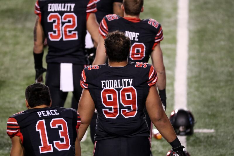 Four members of the Utah Utes football team are seen from the back as they walk across a football field. They are wearing jerseys with words  (“PEACE” “EQUALITY” and “HOPE”) across the back above their numbers.