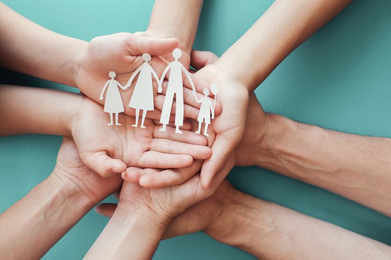 Four sets of hands hold each other up, and the top set of hands holds a paper cutout of a four-person family.