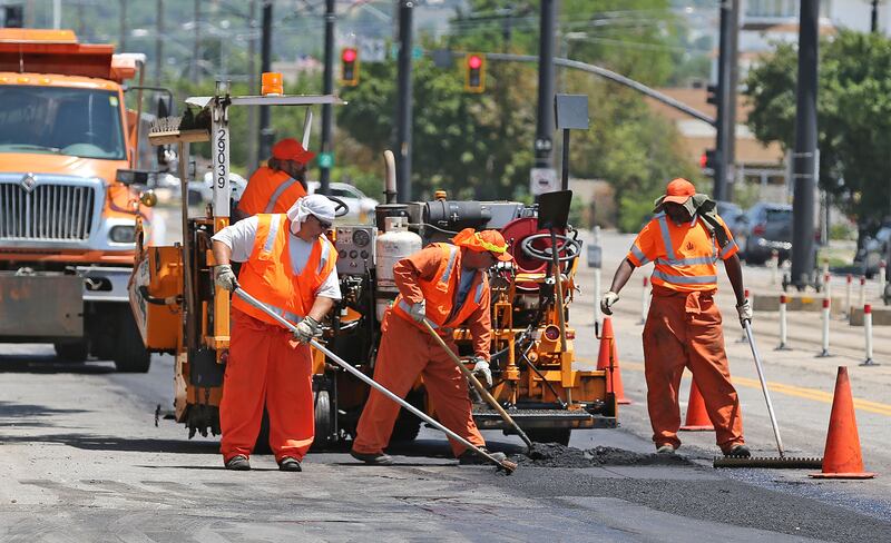 A crew in orange work uniforms works to repave an asphalt road with trucks and orange cones in the background.