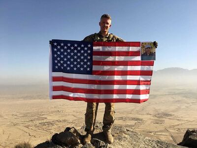 Brent Taylor stands atop the Gharib Ghar peak in Afghanistan while holding an American flag and a photo of John Scharf.