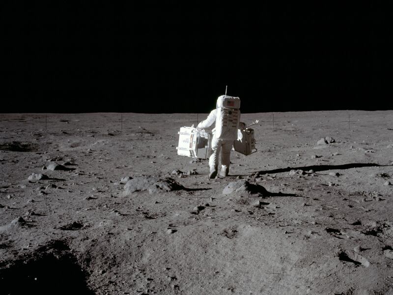 In this July 20, 1969 photo made available by NASA, lunar module pilot Buzz Aldrin carries a seismic experiments package in his left hand and the Laser Ranging Retroreflector to the deployment area on the surface of the moon at Tranquility Base.