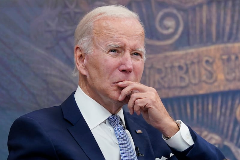 President Biden stands in a navy blue suit with a hand held up to his chin in contemplation.