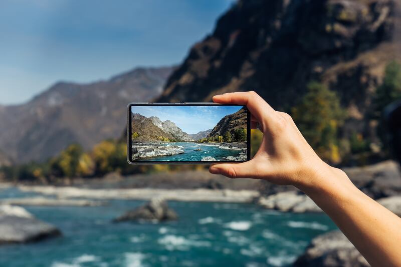 A hand holds a smartphone, taking a photo of mountains and a river.