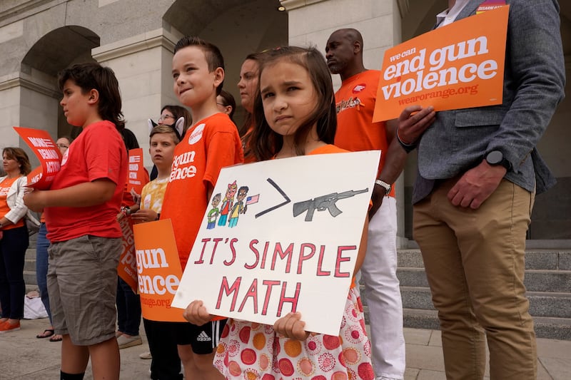 A 7-year-old girl holds a sign with a picture of children, a greater sign, and a semi-automatic rifle with the words “it’s simple math” written below.