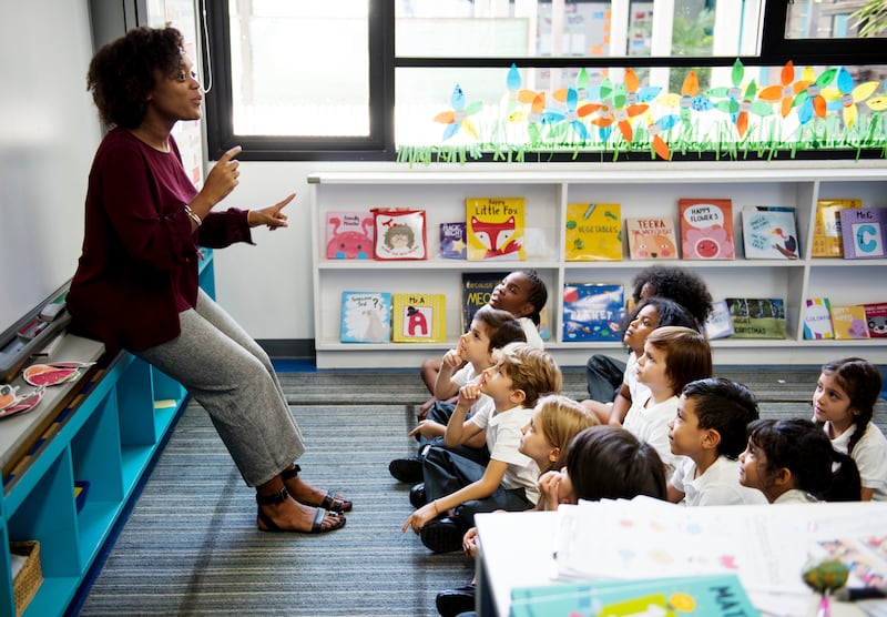 A teacher stands at the front of a classroom addressing a group of small children before her.