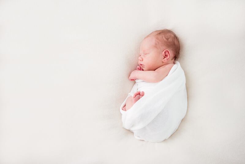 A baby wrapped in a white cloth lays against a cream background.