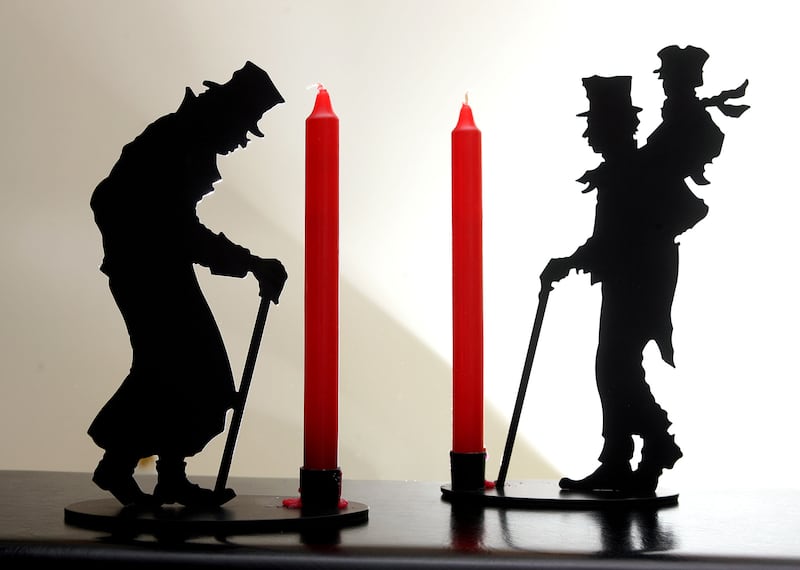 Metal candle holders with red candles and likenesses of Ebenezer Scrooge and Bob Cratchit carrying Tiny Tim sit on a desk.