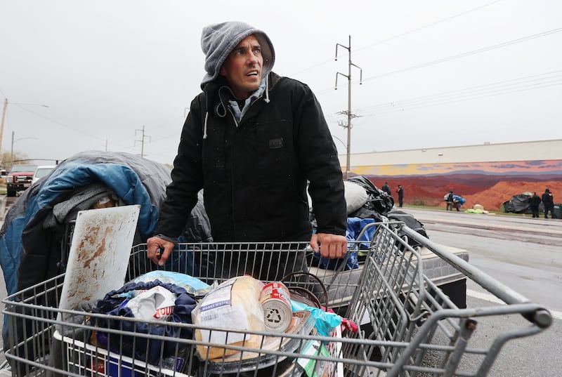 A homeless man stands behind a shopping cart full of belongings on a cloudy day.