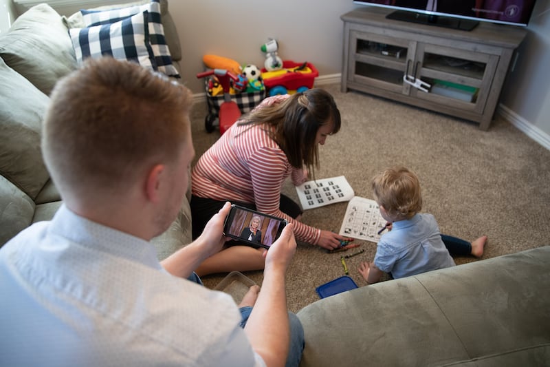 A mother and child play quiet games on the floor while another family member watches a session of general conference.
