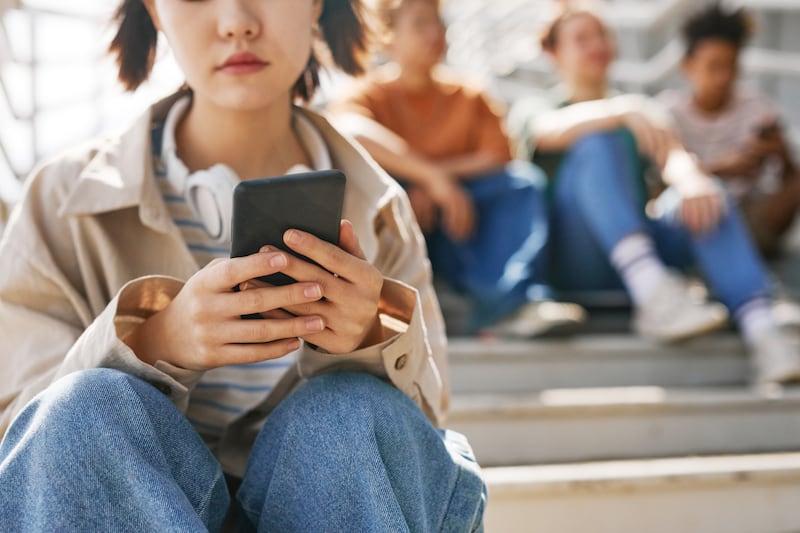 Teenage girl holding smartphone outdoors while sitting on stairs with group of friends in the background.