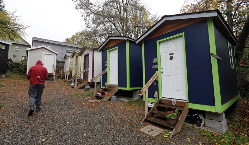 In a file photo from Nov. 9, 2017, a resident walks past a row of tiny houses at a homeless encampment in Seattle where full size homes stand behind. Seattle's quest to create affordable housing now includes restrictions on large houses and greater leeway