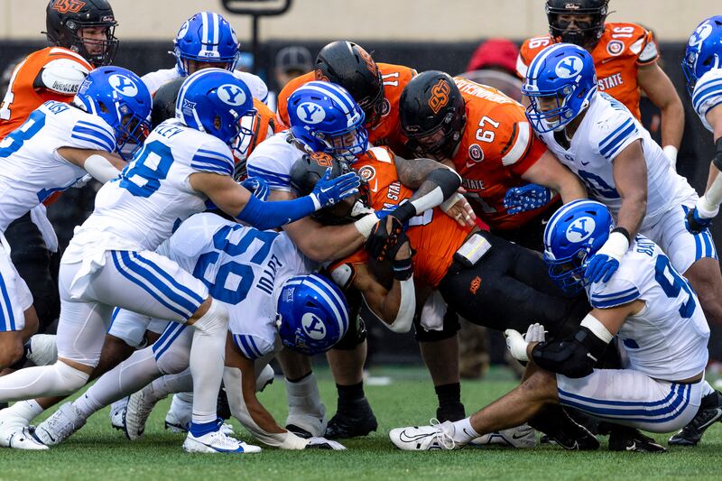 BYU defensive end Logan Lutui, left, defensive tackle Bruce Mitchell, center, and defensive end Tyler Batty, right, tackle Oklahoma State running back Ollie Gordon II (0) in the first half of an NCAA college football game Saturday, Nov. 25, 2023, in Stillwater, Okla.