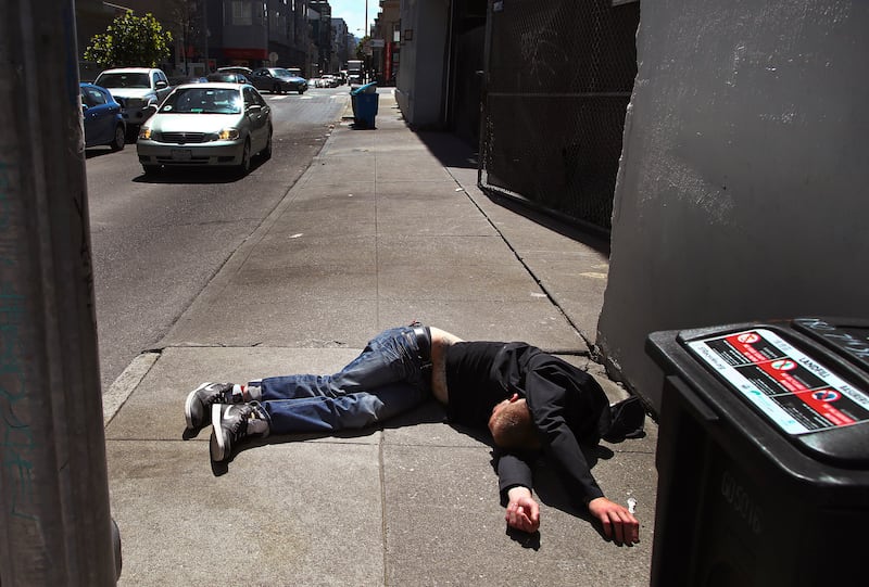 A man lies on the sidewalk beside a recyclable trash bin in San Francisco.