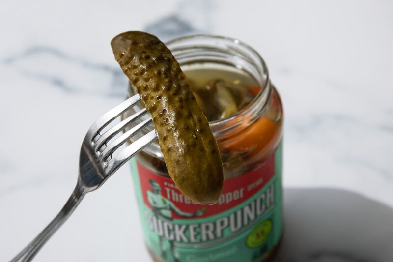 A stainless steel fork lifts a pickle out of an open pickle jar placed on a white marble counter.