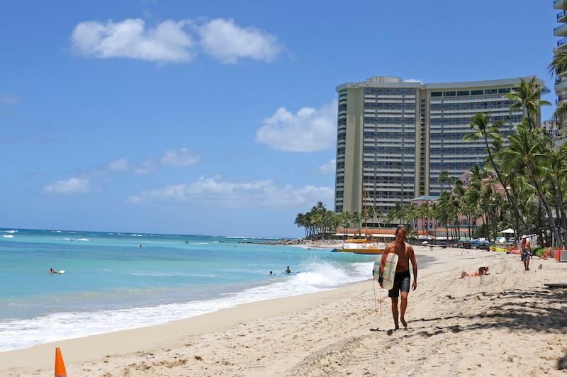 In this photo taken Friday, June 5, 2020, a surfer walks on a sparsely populated Waikiki beach in Honolulu. Hawaii faces unpleasant options for addressing a dramatic decline in tax revenues precipitated by the coronavirus pandemic and the shutdown of the state’s tourism industry.