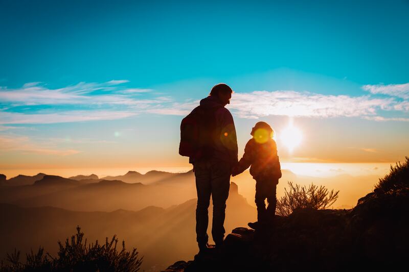 A father and daughter stand silhouetted on a hill overlooking the sunrise.