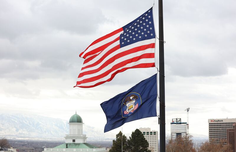 The Utah state flag with the symbolic beehive and the American flag of starts and stripes wave in the sky on a cloudy day.