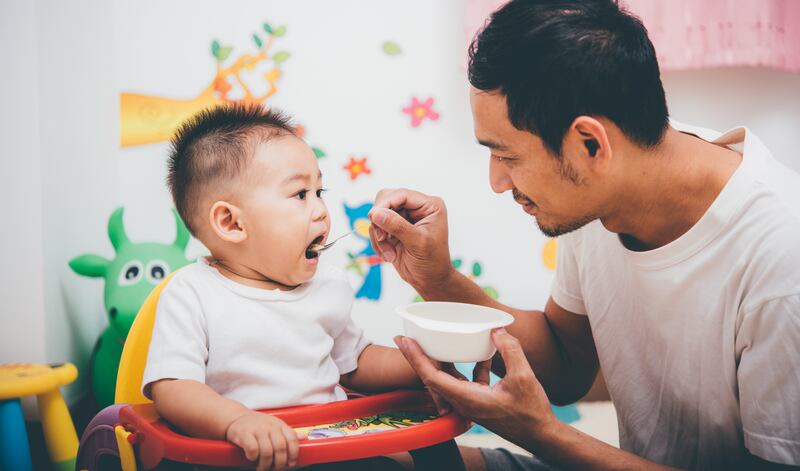 A father spoons food into the wide open mouth of a 1-year-old baby in a yellow and red high chair.