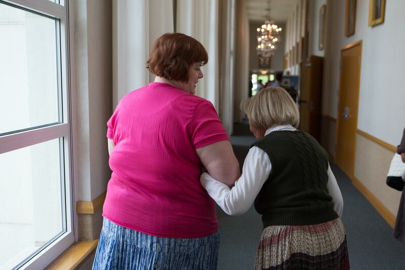 A woman helps an elderly woman walk down a hallway.