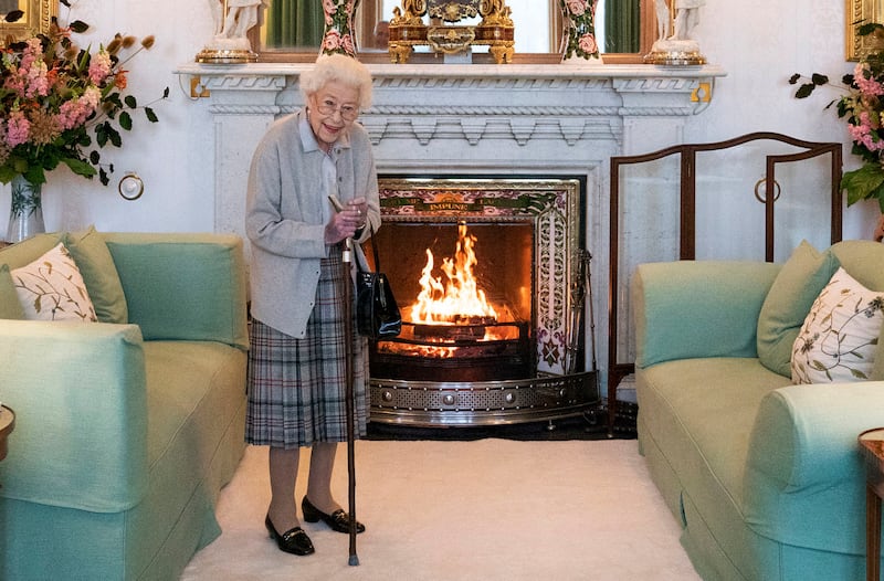 Queen Elizabeth II, smiling, stands with a cane in a drawing room with a lit fire and two mint green couches.