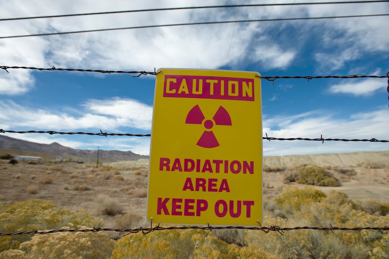 A yellow sign attached to a barbed-wire fence reads “CAUTION. RADIATION AREA. KEEP OUT” in bold red letters.