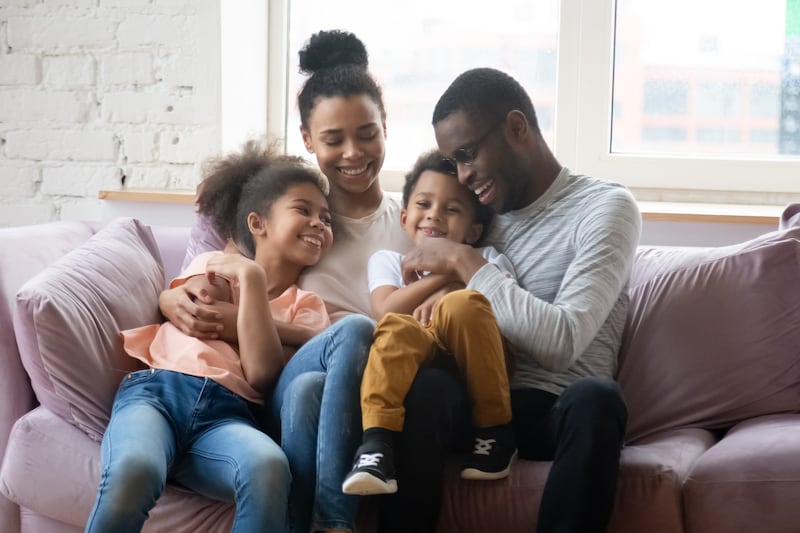 A smiling family of four sits on a couch and the parents hug the two children.