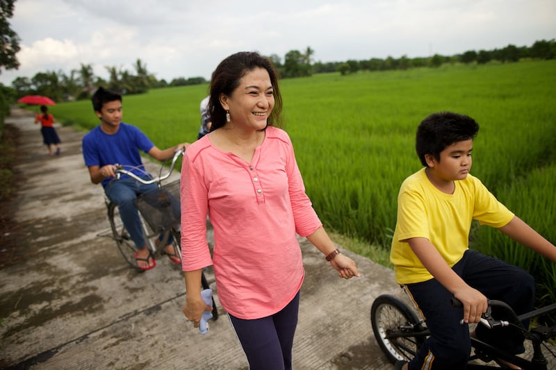 A mother walks next to her two sons, who are riding their bicycles on a path next to a field of green plants.