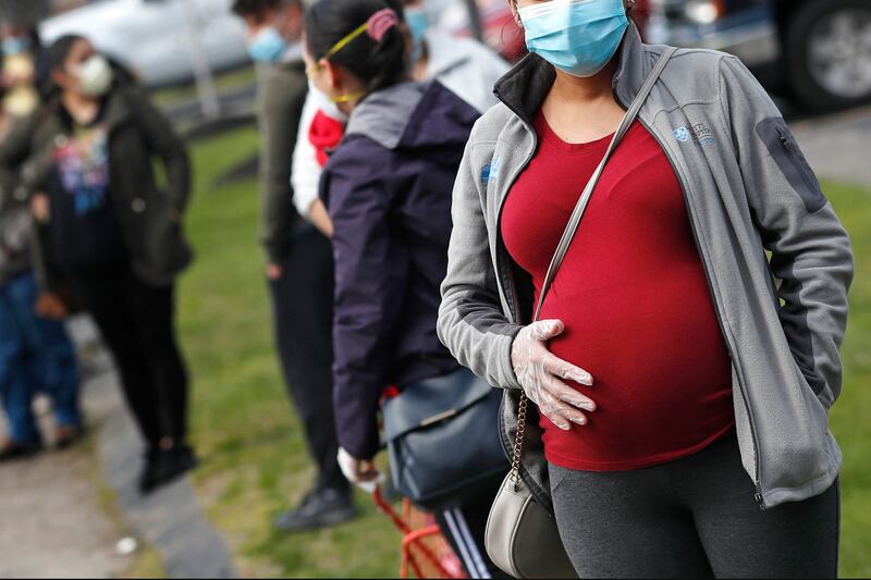 In this Thursday, May 7, 2020 file photo, a pregnant woman wearing a face mask and gloves holds her belly as she waits in line for groceries with hundreds during a food pantry sponsored by Healthy Waltham for those in need due to the COVID-19 virus outbreak, at St. Mary’s Church in Waltham, Mass.