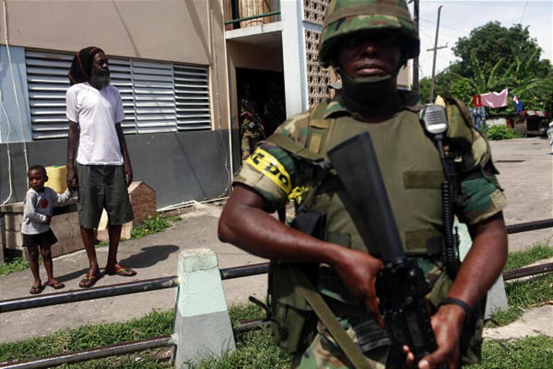 A man watches as he holds his son's hand while a soldier stands guard in Tivoli Gardens in Kingston, Jamaica.