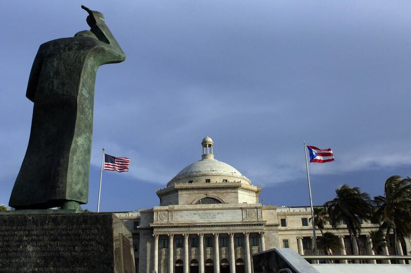 FILE - In this Wednesday, July 29, 2015, file photo, a bronze statue of San Juan Bautista stands in front of Puerto Rico’s Capitol as U.S. and Puerto Rican flags fly in San Juan, Puerto Rico. After months of pleading from the government of Puerto Rico, th