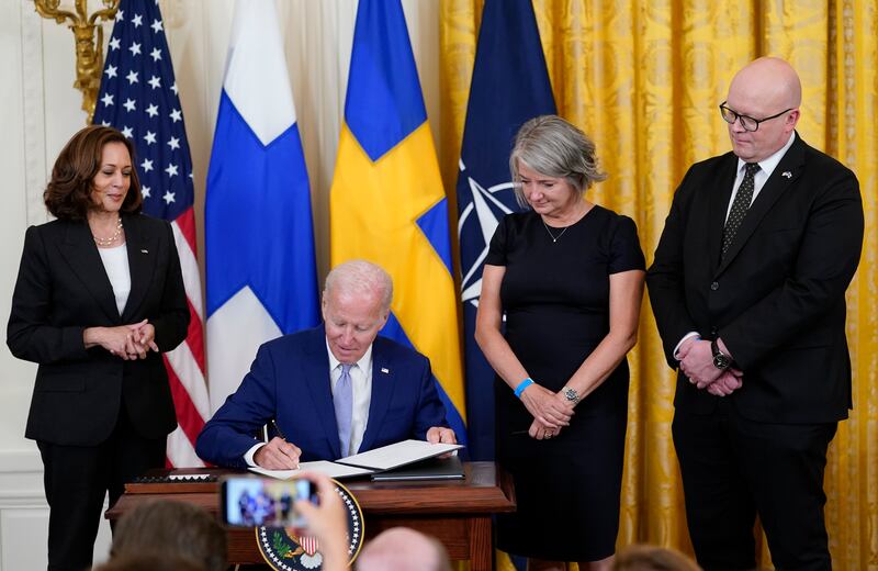 President Joe Biden signs a treaty in front of several country flags, Kamala Harris, Karin Olofsdotter, Sweden’s ambassador to the U.S., and Mikko Hautala, Finland’s ambassador to the U.S.