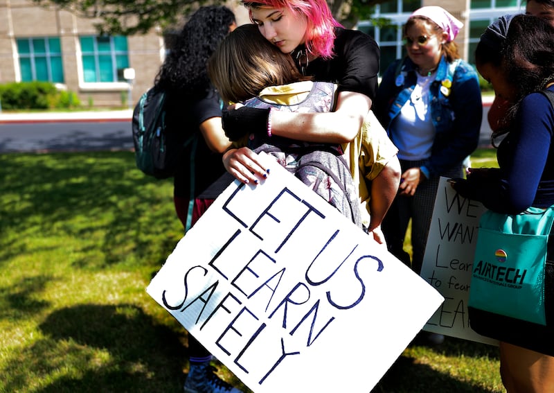 Two high schoolers hug in front of a school while one holds a sign that reads “Let us learn safely.”
